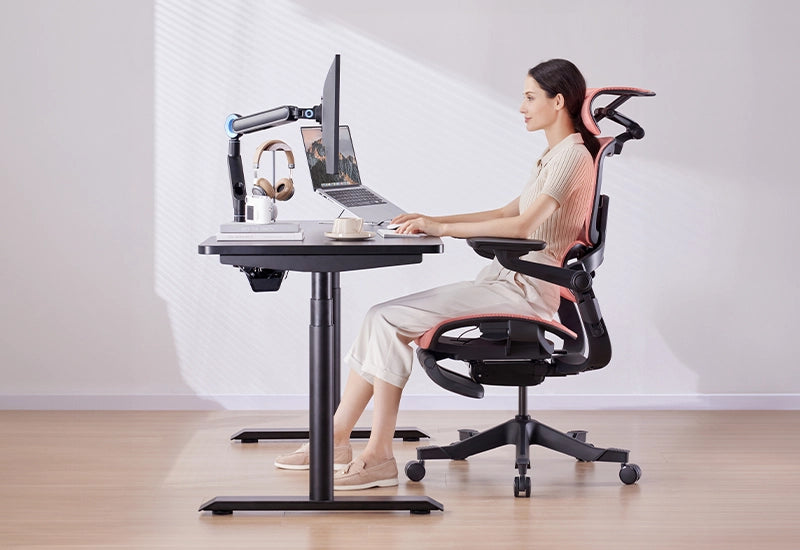 Woman using a H1 Pro ergonomic office chair at a height-adjustable desk with dual monitor setup in a bright workspace.